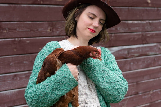 una chica del campo sostiene un pollo en sus manos, se para cerca del granero
 - Foto, imagen