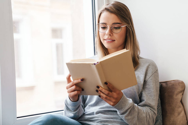 Photo of serious beautiful woman in eyeglasses reading book while sitting near window indoor - Foto, imagen