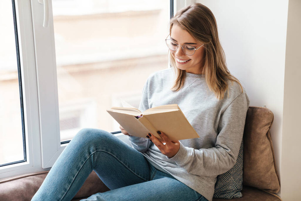 Photo of smiling beautiful woman in eyeglasses reading book while sitting near window indoor - Foto, immagini