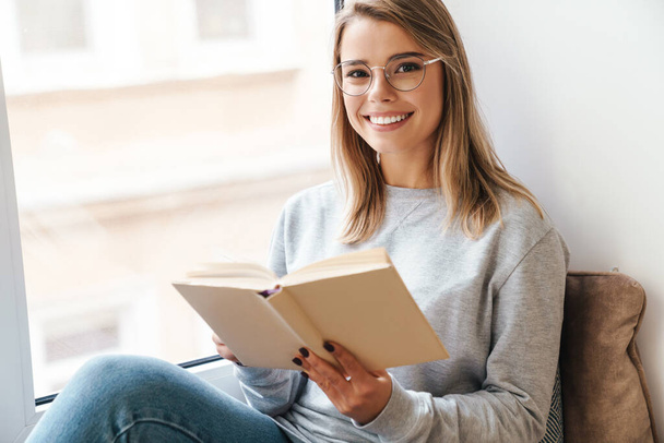 Photo of smiling beautiful woman in eyeglasses reading book while sitting near window indoor - Foto, afbeelding