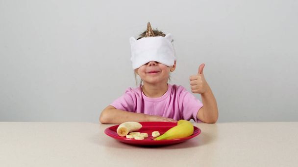 Klein meisje in een masker probeert en raadt banaan en geeft duimen omhoog - Foto, afbeelding