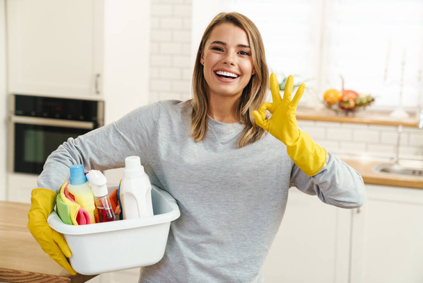 Photo of smiling young woman housewife in gloves holding cleanser bottles and showing ok sign at modern kitchen - Valokuva, kuva