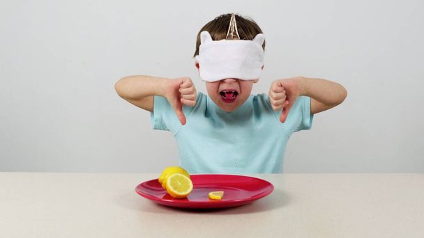Kleiner Junge in Maske versucht Zitrone zu erraten und drückt Daumen nach unten - Foto, Bild