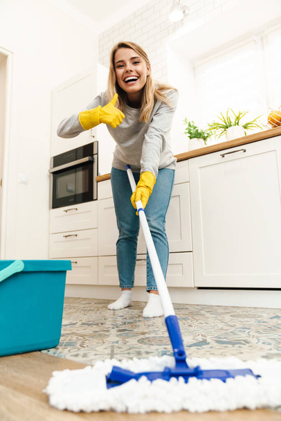 Photo of joyful young woman housewife in gloves gesturing thumb up and winking while mopping floor at kitchen - 写真・画像