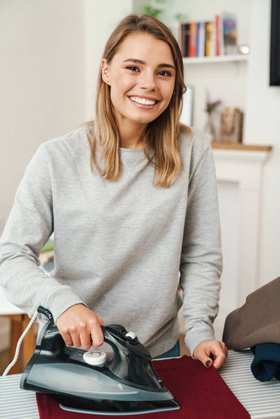 Photo of cheerful young beautiful woman housewife smiling while ironing clothes at living room - Fotoğraf, Görsel