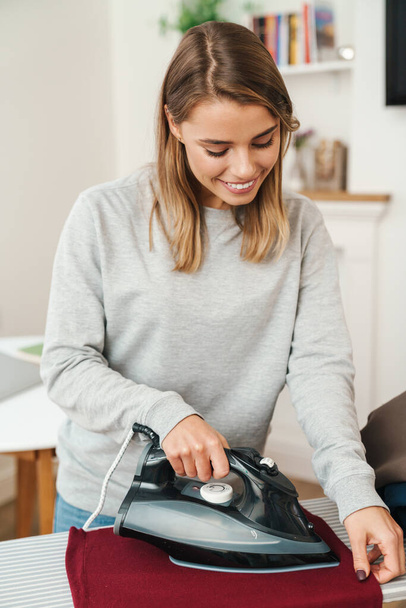 Photo of cheerful young beautiful woman housewife smiling while ironing clothes at living room - Valokuva, kuva