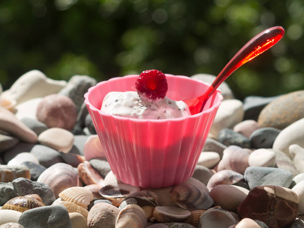 Розовая миска белого мороженого с красной малиной и маленькой пластиковой ложкой на пляжных камнях при солнечном свете
 - Фото, изображение