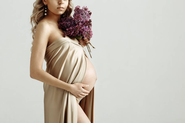 Close up of pregnant woman holding a bouquet of lilac flowers and touching her belly on gray background, isolated. Pregnancy, maternity, preparation and expectation concept - Photo, Image