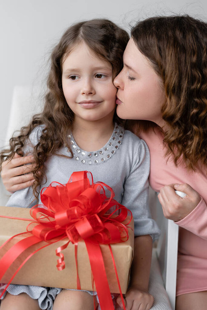 Ünnepnap. Egy 6-7 éves lány ül egy székben. Hullámos haja van. Anya adott neki egy ajándékot egy gyönyörű dobozban, piros masnival. A szüleik boldog gyermeke. Ünnepi koncepció. - Fotó, kép