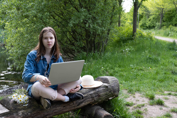 near the lake, outside the city, a young girl sits on an old log with a laptop, in spring - Φωτογραφία, εικόνα