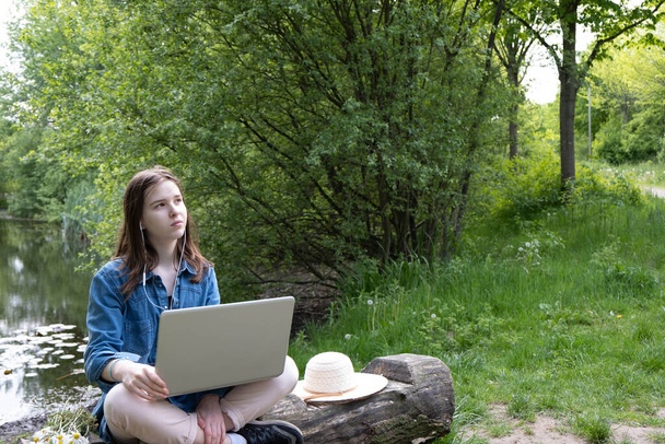 cerca del lago, fuera de la ciudad, una joven se sienta en un viejo tronco con una computadora portátil y mira al cielo en primavera
 - Foto, imagen