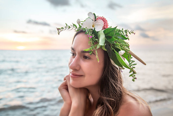 Mooie jonge vrouw met een krans van bloemen op haar hoofd door de oceaan bij zonsondergang. - Foto, afbeelding