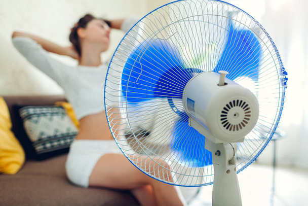 Summer heat. Air conditioning. Young woman cooling down sitting on couch by ventilator at home. Modern devices. - Zdjęcie, obraz