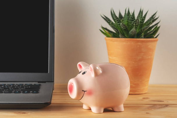 Piggy bank on a wooden table with a laptop. Saving money concept. - Zdjęcie, obraz