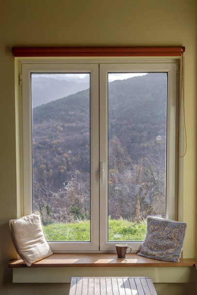 Vue de la fenêtre fermée sur les montagnes et une tasse de café sur le rebord de la fenêtre par une journée ensoleillée d'hiver - Photo, image