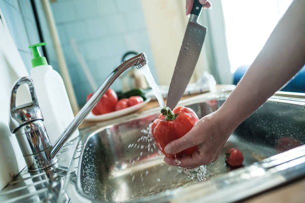 La mano femenina sostiene el pimiento rojo bajo un chorro de agua y lo va a cortar con un cuchillo, en la cocina. Cocinar alimentos
. - Foto, Imagen