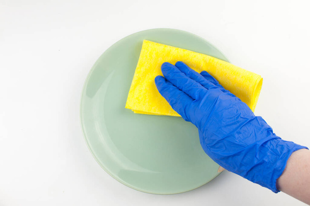 Un plato verde vacío en las manos de guantes azules con un trapo amarillo sobre un fondo blanco
. - Foto, imagen