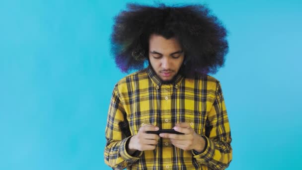 Afroamerikanischer Mann spielt Spiel auf Smartphone auf blauem Hintergrund. Konzept der Emotionen - Filmmaterial, Video