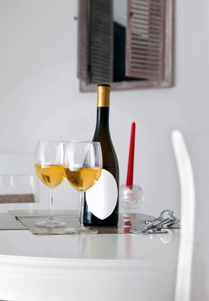 Láhev vína s bílým štítkem na titulek, otvírák, dvě sklenice bílého vína na stůl a okno na pozadí - Fotografie, Obrázek
