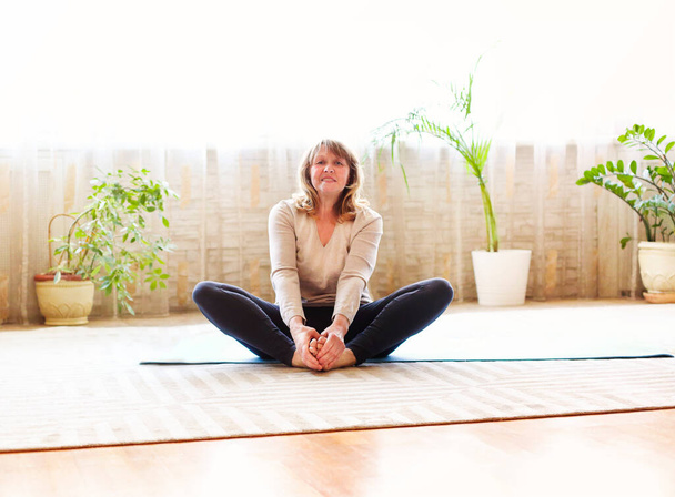 Corpo inteiro descalço idosa sentada no chão em postura de borboleta e alongamento pernas enquanto faz ioga em quarto espaçoso em casa
 - Foto, Imagem