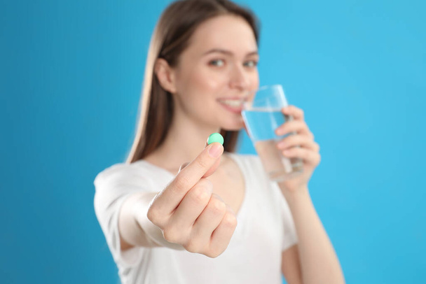 Young woman with glass of water and vitamin pill against blue background, focus on hand - Fotoğraf, Görsel