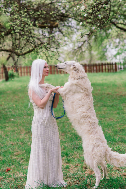 Blonde woman in white dress playing with her Russian wolfhound in the garden - Photo, Image