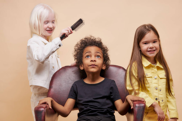 multiracial kid boy sit on chair as a king while his friends comb his hair - Fotó, kép