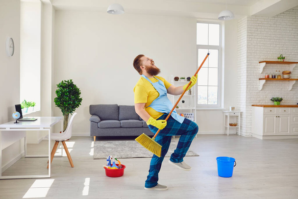 Funny fat man with a beard in an apron dancing cleans the room in the house. - Foto, Imagen