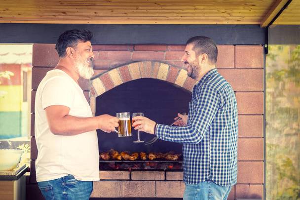 Çok ırklı erkek arkadaşlar barbekü ızgarasında et pişirirken alkol içerek eğleniyorlar. - Fotoğraf, Görsel