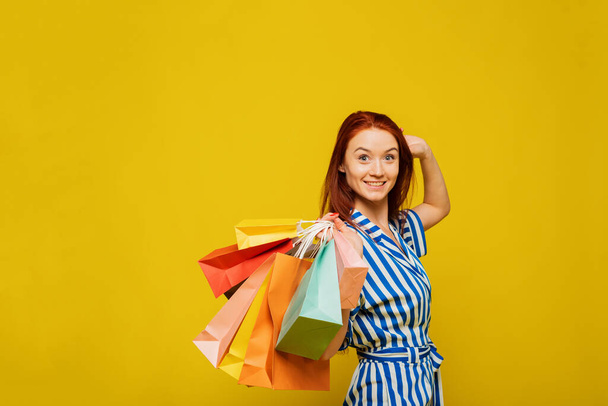 Das rothaarige Mädchen hält Pakete mit Einkäufen auf gelbem Hintergrund. Konzept des Einkaufs und Verkaufs von glücklichen jungen Mädchen mit Paketen. - Foto, Bild