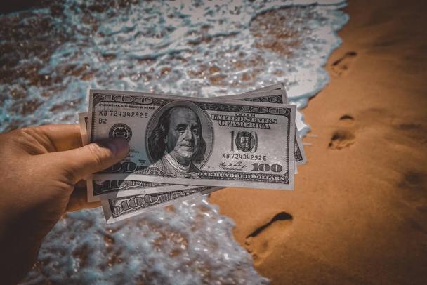 Girl holding money bill of 300 dollars on background of sea ocean waves with white foam and sand wet beach with footprints in sand close-up. Concept finance money holiday traveling dollars vacation - Foto, imagen