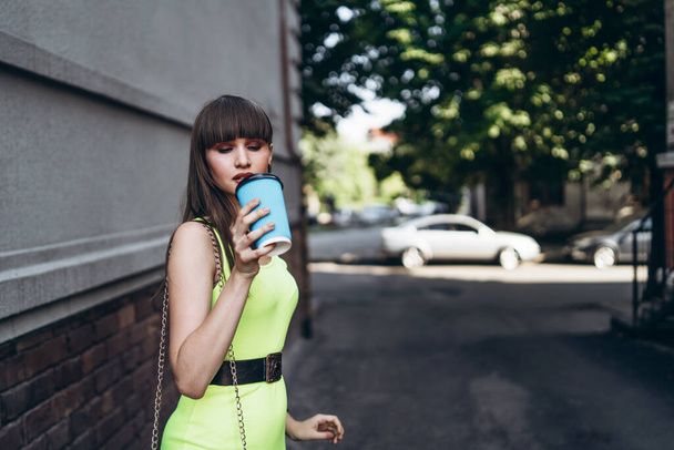 Pretty long hair brunette girl in green light dress with blue paper cup of beverage walking outdoor on the street in the city - Zdjęcie, obraz