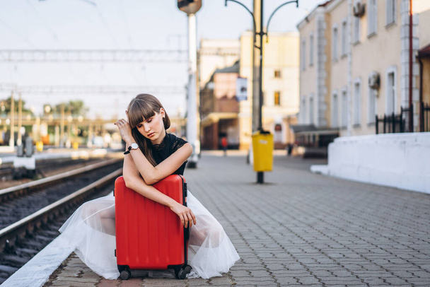 Γυναίκα μελαχρινή ταξιδιώτισσα με κόκκινη βαλίτσα σε λευκή φούστα περιμένει για ένα τρένο στο σταθμό raiway - Φωτογραφία, εικόνα