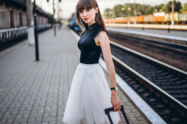 Female brunette traveler with red suitcase in white skirt waiting for a train and walking on raiway station - Fotografie, Obrázek