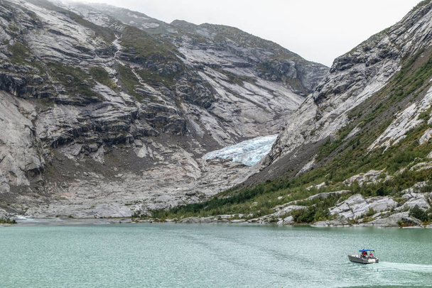 Noruega montañas paisaje vista cerca del lago con agua azul azur, barco, rocas, glaciar, nieve. Viaje de día nublado a Nigardsbreen en el parque nacional Jostedalsbreen
 - Foto, imagen