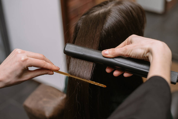 The hairdresser pulls and straightens the hair of a brunette in a beauty salon with a comb and ironing. Keratin recovery hair and protein treatment pile with professional ultrasonic iron tool. - Foto, Imagen