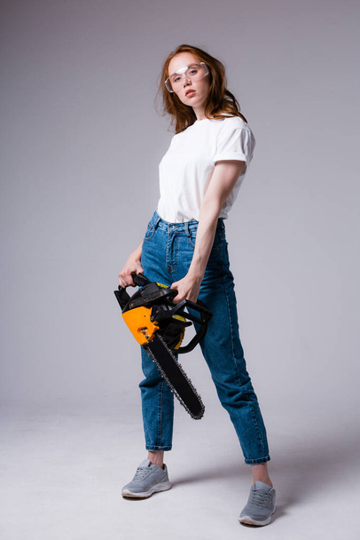 A girl with a chainsaw in her hands and in safety glasses stands half a turn and looks at the camera. - Foto, Bild