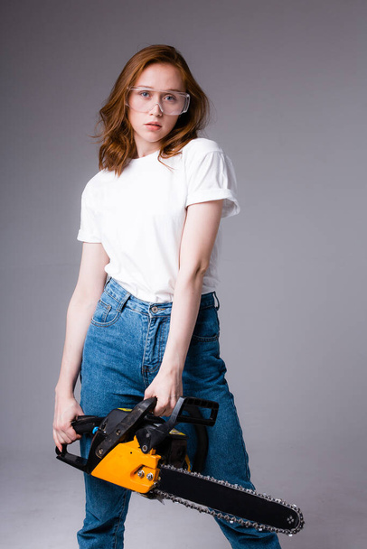 Red-haired girl in protective glasses holds chainsaw in her hands and looks at camera. Photo for poster of construction company - Foto, Imagen
