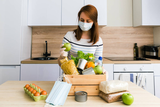 Beautiful girl in medical gloves, mask dial a charity donation crate food box: oil, water, herbs, apples and fruits, cereals canned food, rice, pasta, medical masks. White wooden kitchen background - Foto, immagini