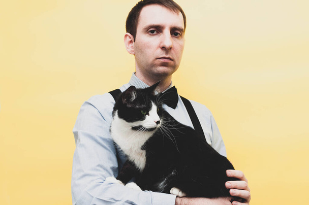handsome confident man in blue shirt holding black and white taxedo cat and looking at camera in front of yellow background - Foto, Imagem