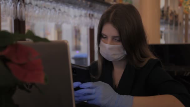 Mädchen slawische Erscheinung in medizinischen Maske und Gummihandschuhen funktioniert Computer-Laptop. Frau schaut aufs Telefon, lächelt, denkt nach und setzt Maske auf. Junge Frau in Großaufnahme. Konzept funktioniert während der Covid-19-Pandemie - Filmmaterial, Video