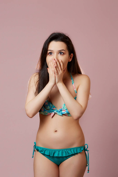 a young beautiful slender Asian girl in a swimsuit covered her mouth with her hand in surprise, in admiration, in delight .  on a pink isolated background. - Photo, Image