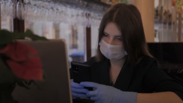 Girl Slavic appearance in medical mask and rubber gloves works computer-laptop. Woman looks at phone, smiles, thinks, and puts on mask. Close-up young woman. Concept working during Covid-19 pandemic - Video