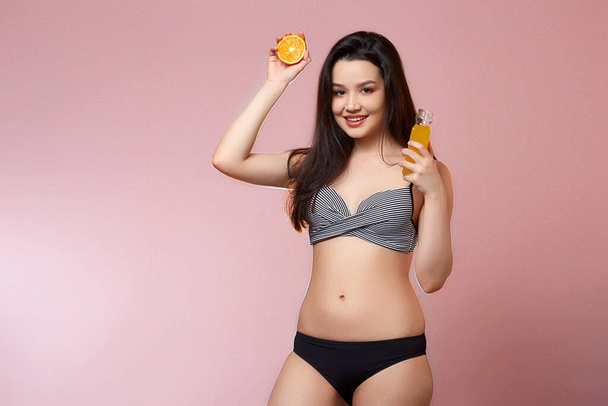 young beautiful slender Asian girl in a swimsuit . with fresh juice and an orange in his hands. smiles. on a pink isolated background. - Photo, Image