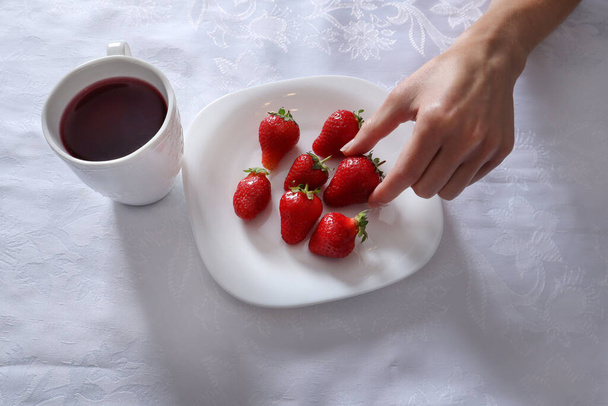 Mano de niña toma fresas del plato. Un plato está lleno de fresas y una taza de té en un mantel blanco. Comida de temporada y concepto de fruta fresca. Primer plano, enfoque selectivo
 - Foto, Imagen