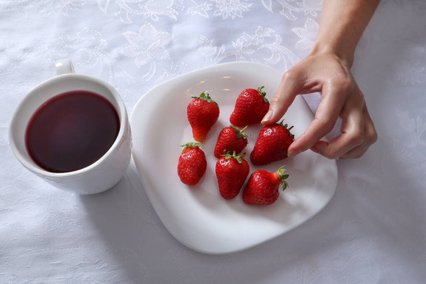 Vrouwenhand neemt aardbeien van de plaat. Een bord staat vol aardbeien en een kopje thee op een wit tafelkleed. Seizoensgebonden voedsel en vers fruit concept. Close-up, selectieve focus - Foto, afbeelding