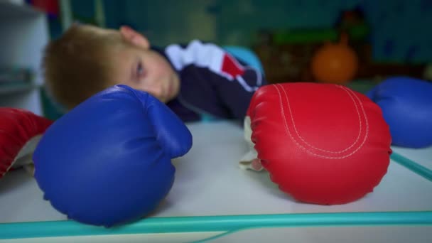 An upset child looks out the window during quarantine. Sad boy in boxing gloves is sitting at the table. The boy dreams of playing box with friends. Longing for the game in the fresh air. Loneliness.  - Footage, Video