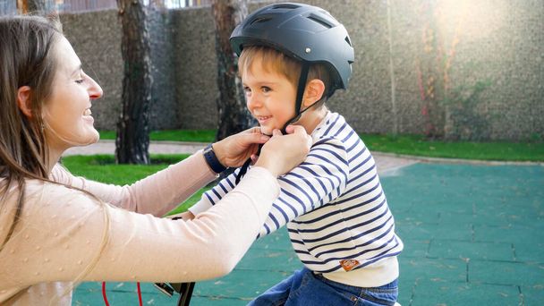 笑顔の若い母親の肖像自転車に乗る前に彼女の息子保護ヘルメットバックルを調整し、固定 - 写真・画像