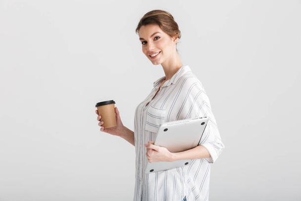 Obraz radosnej pięknej kobiety pijącej kawę na wynos i trzymającej laptopa na białym tle - Zdjęcie, obraz