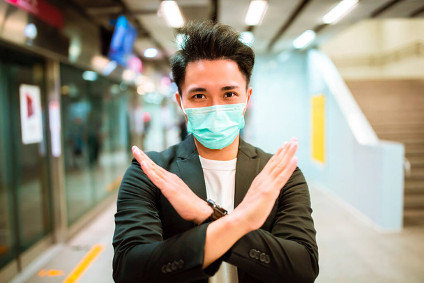 νεαρός επιχειρηματίας φορώντας μάσκα προσώπου και δείχνει στοπ χειρονομία πινακίδα στο σιδηροδρομικό σταθμό  - Φωτογραφία, εικόνα